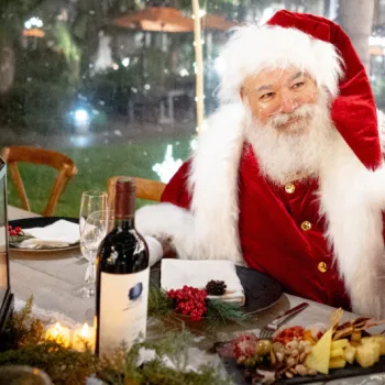 Santa Claus sits at a festive table with wine, food, and holiday decorations, illuminated by candlelight and outdoor lights in the background.