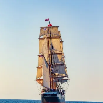 A tall ship with multiple sails unfurled sails on open water under a clear sky, flying a flag at the top of its mast.
