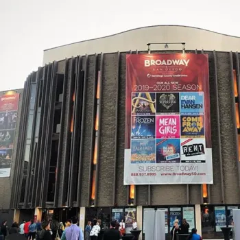 A theater building with a large banner displaying Broadway San Diego’s 2019-2020 season lineup, while people gather outside the entrance.