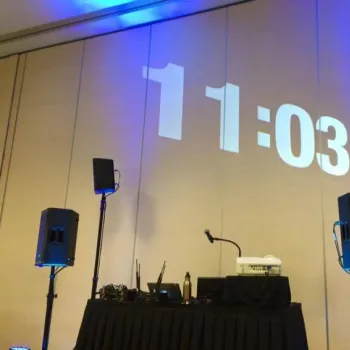 A digital clock displaying 11:03 is projected onto a wall above a table with electronic equipment and speakers in a room with blue lighting.