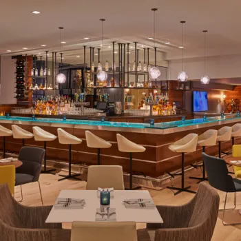 Modern bar and lounge area with a central bar surrounded by white barstools, tables with chairs, pendant lights, and assorted bottles on display.