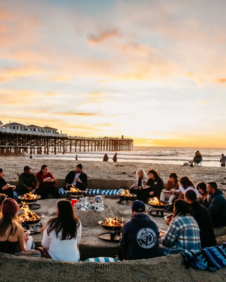A group of people sit around fire pits in a sand pit on the beach at sunset, with a pier and ocean in the background.