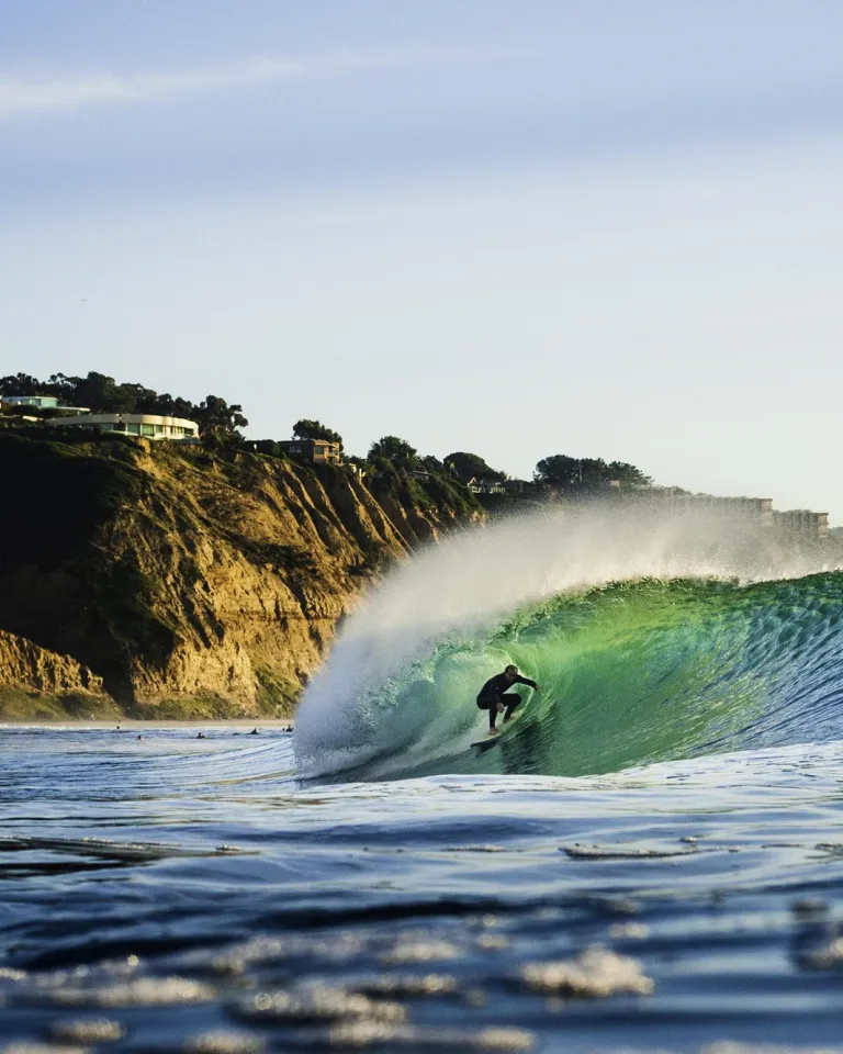 A surfer rides inside the barrel of a wave near a rocky coastline with cliffs and houses visible in the background.