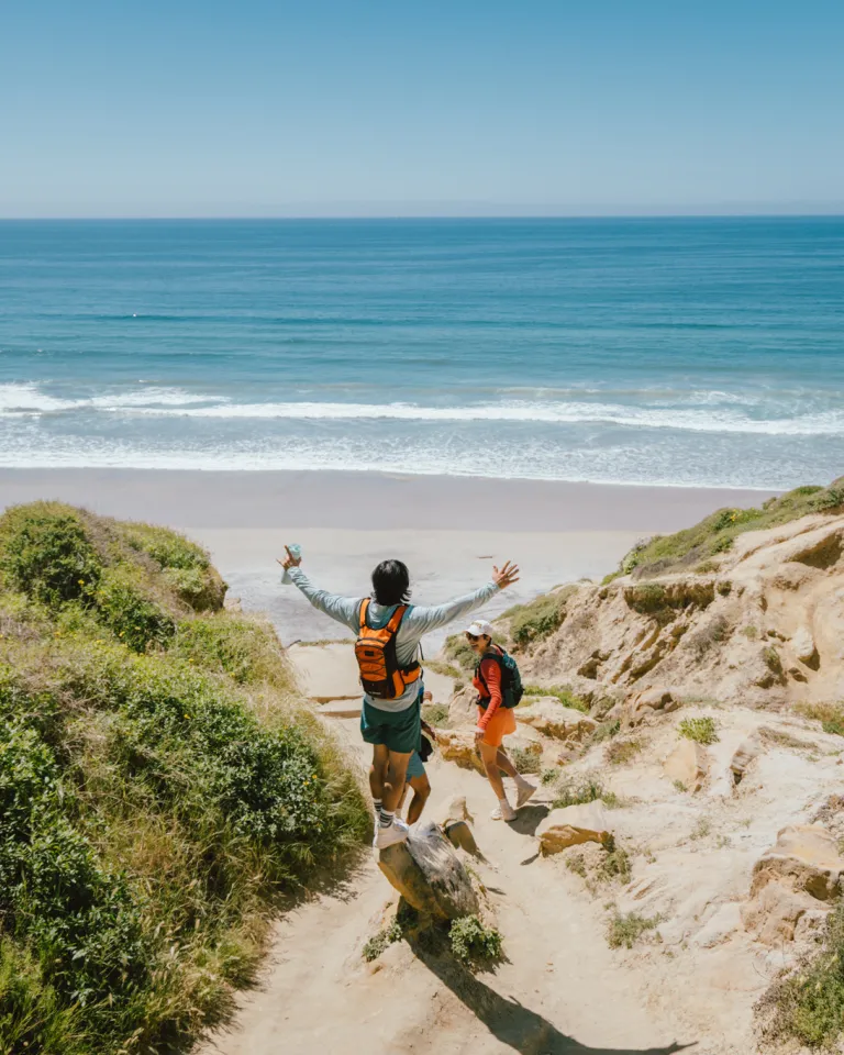 Two people with backpacks walk down a sandy trail in Torrey Pines toward a Blacks Beach, surrounded by greenery and cliffs, with the ocean visible in the background.