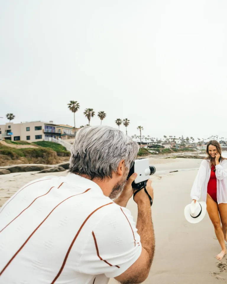 A man takes a photo of a woman posing on a sandy San Diego beach near the ocean. The woman holds a hat and smiles, and palm trees and houses are visible in the background.