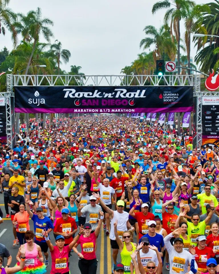 Large crowd of runners in colorful outfits starts the Rock 'n' Roll San Diego Marathon, with palm trees and banners visible in the background.