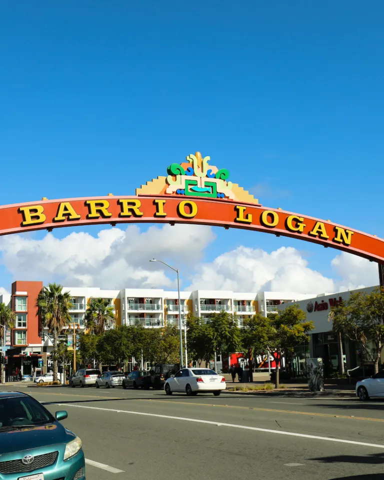 A large red arch with "Barrio Logan" written on it spans a street lined with parked cars and buildings under a blue sky.