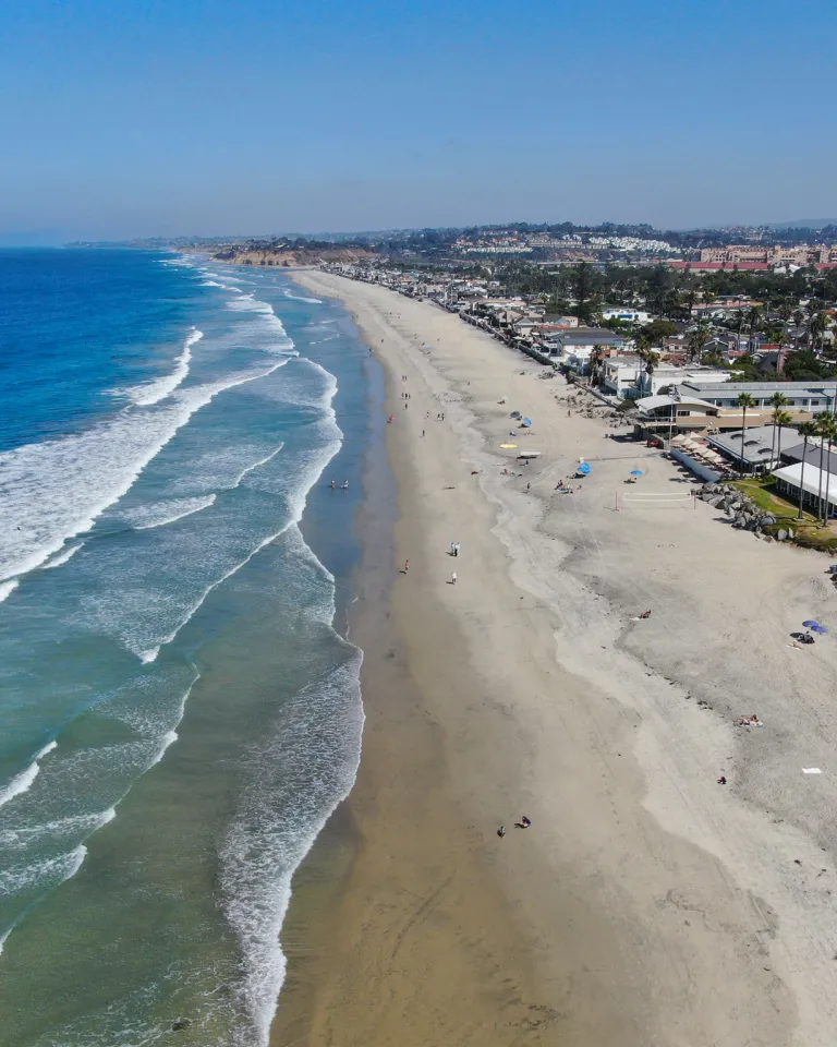 Aerial view of Del Mar's wide sandy beach with waves along the shoreline, people scattered on the sand, and buildings lining the coast under a clear blue sky.