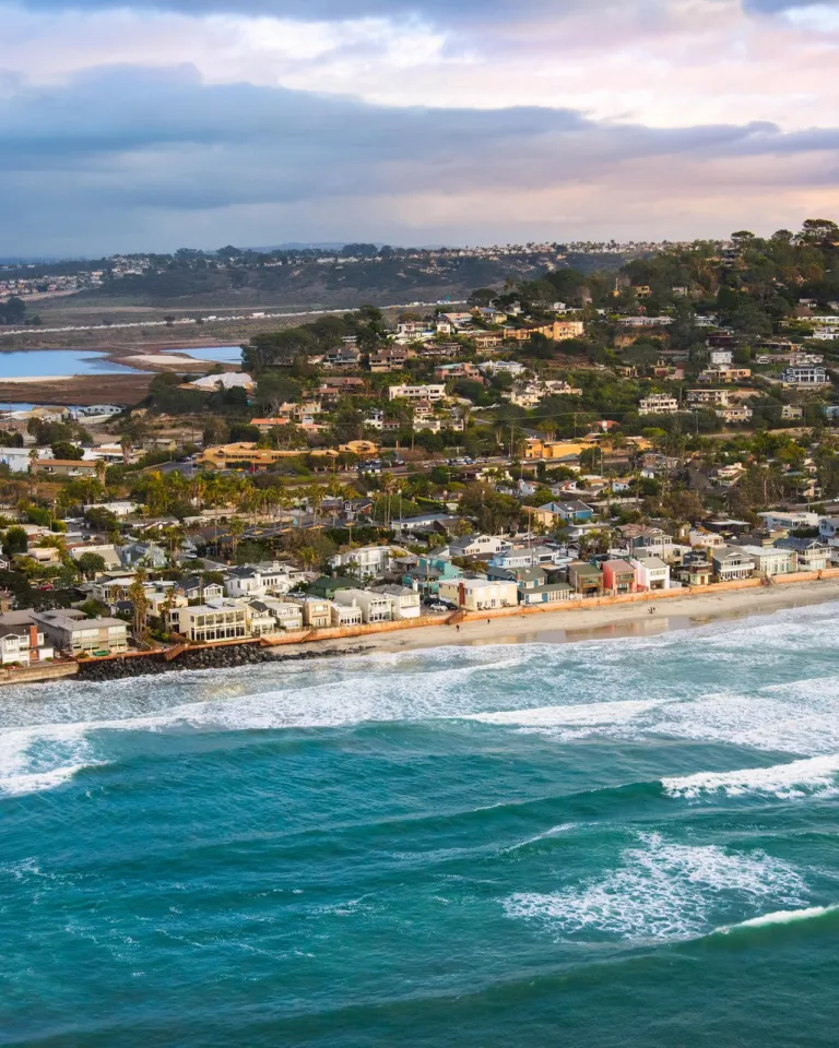 A coastal town with houses lined along the shore, waves crashing on the beach, and rolling hills in the background under a partly cloudy sky.