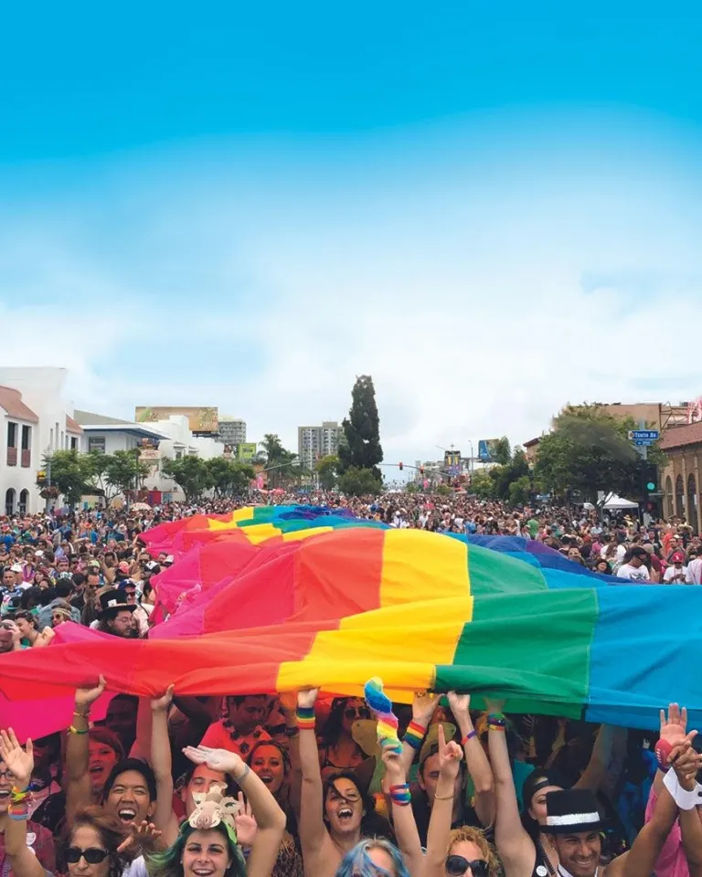 Large crowd of people at a street parade holding up a giant rainbow flag, with buildings lining both sides and a clear blue sky above.