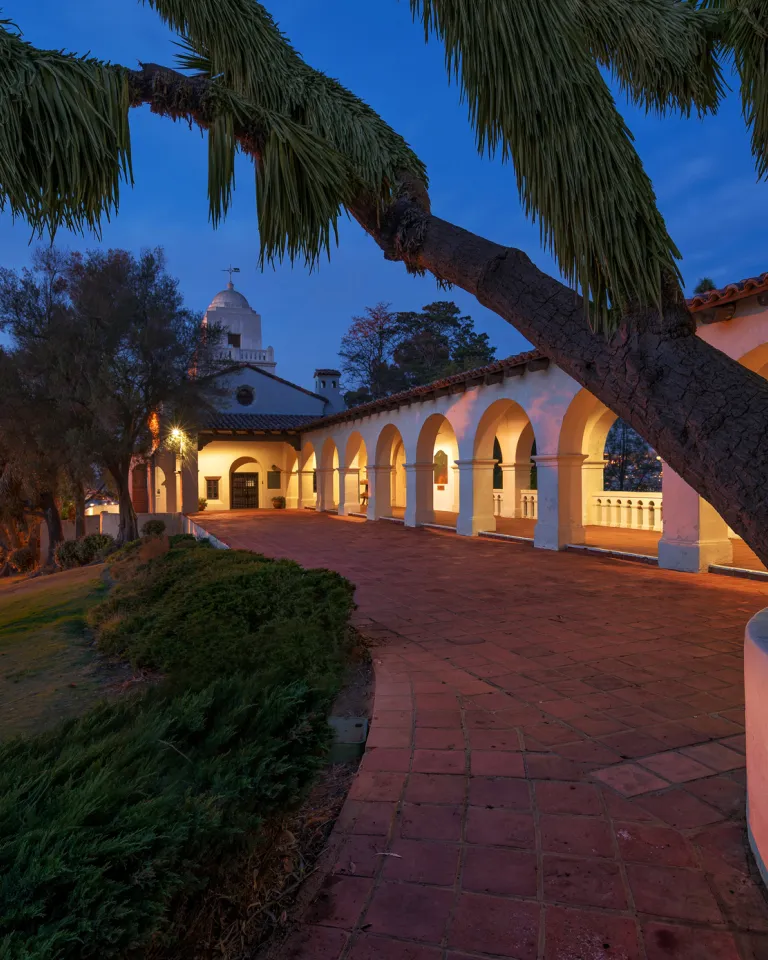 A large tree frames a Spanish-stylePresidio Museum with arches and a dome, illuminated at dusk, overlooking a cityscape with visible lights in the distance.