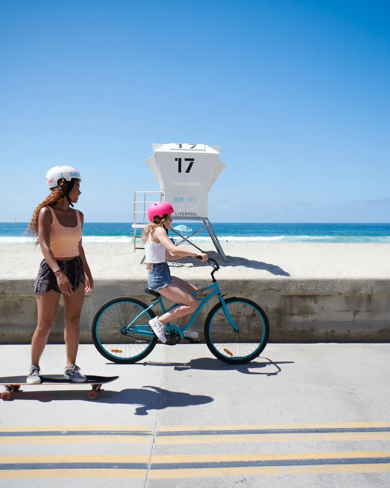 Two teenagers on a bike and skateboard cruising down the Board Walk of Mission Beach in San Diego
