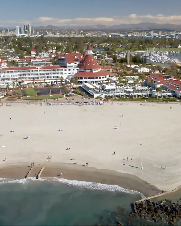 Aerial of Coronado Beach with the Hotel Del Coronado, Coronado Bay Bridge, and San Diego Skyline in the Background.