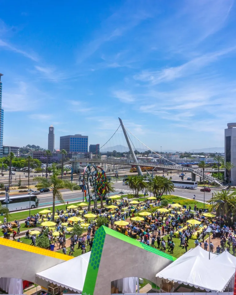 Outdoor event with crowds, yellow umbrellas, palm trees, and a Ferris wheel; city buildings, a bridge, and blue sky in the background.