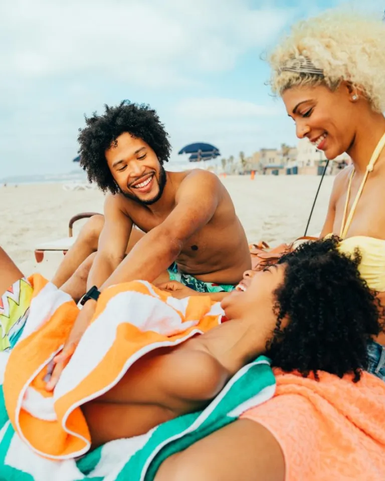 Three people sit smiling and laughing together on a beach, sitting on towels under a canopy with sand and ocean visible in the background.