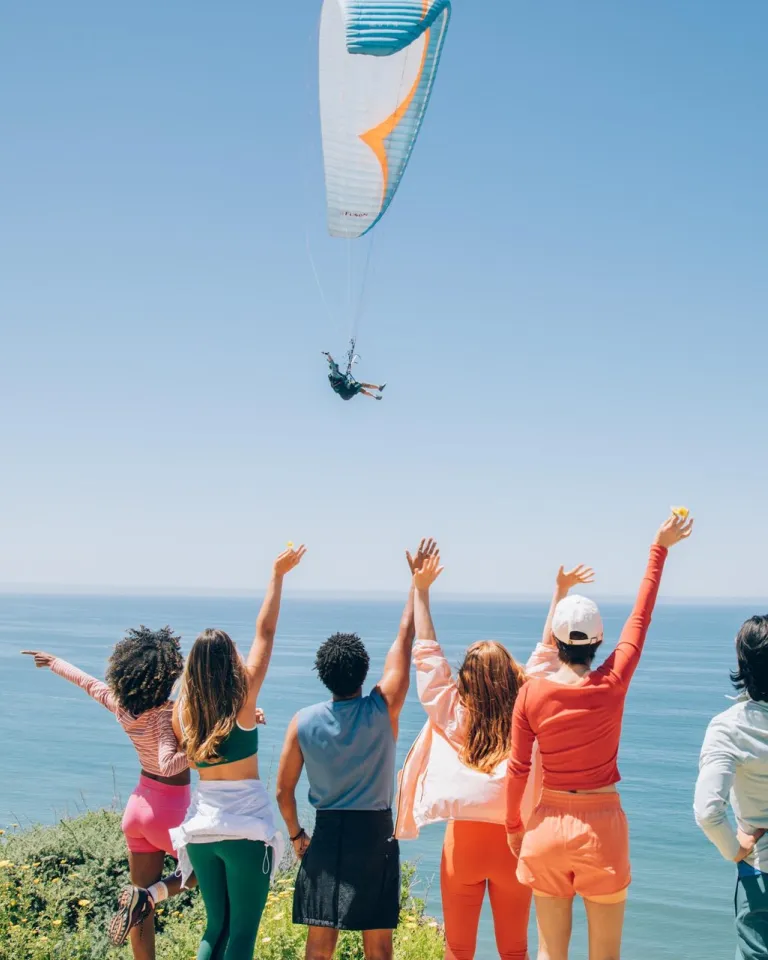 Six people stand on a grassy bluff at the Torrey Pines Gliderport in the La Jolla neighborhood of San Diego, facing the ocean with arms raised as they watch a paraglider flying over the water under a clear blue sky.