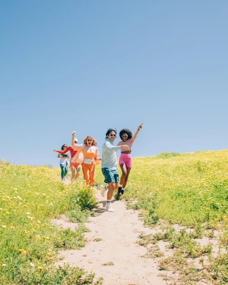 Five people run down a dirt path through a field of yellow wildflowers under a clear blue sky on a seaside bluff in San Diego.