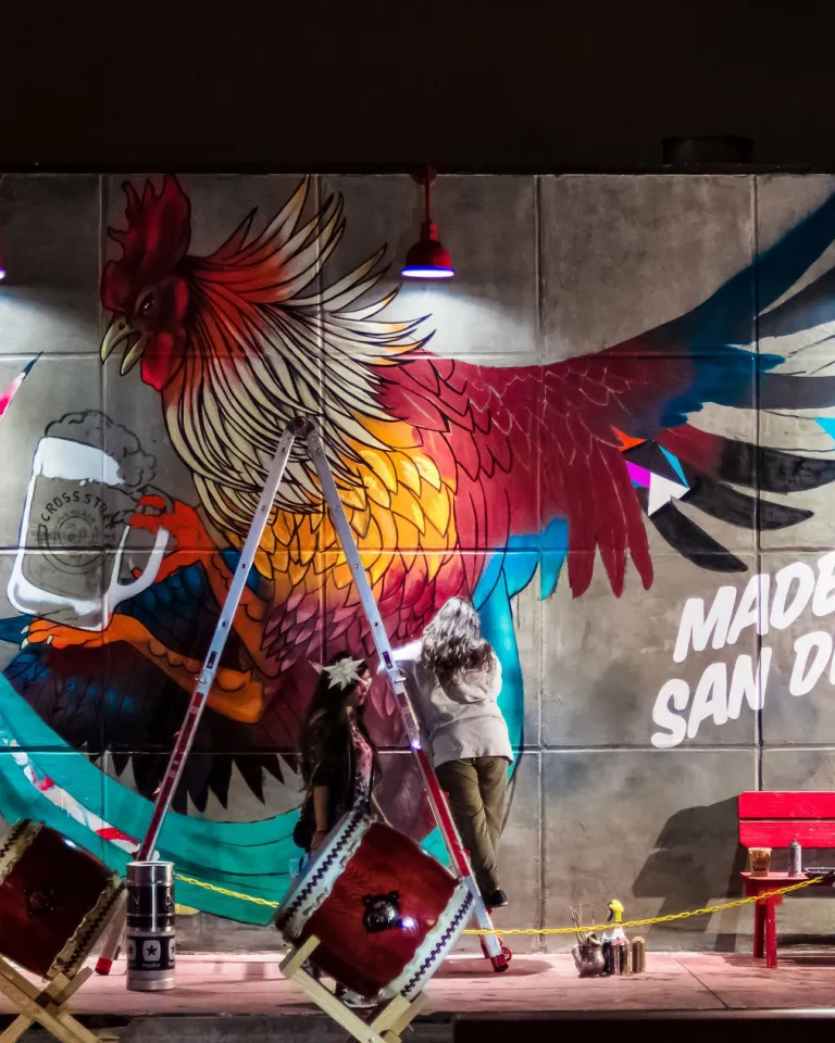 A mural of a colorful rooster holding a beer mug with the text "Made in San Diego" painted on a concrete wall at night; red bench and drums are in front.