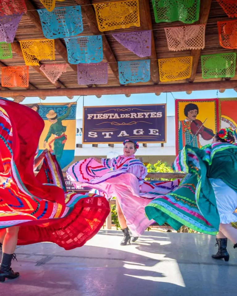 Three dancers in colorful traditional dresses perform on an outdoor stage at Fiesta de Reyes in San Diego's Old Town, decorated with papel picado banners and Fiesta de Reyes signs in the background.