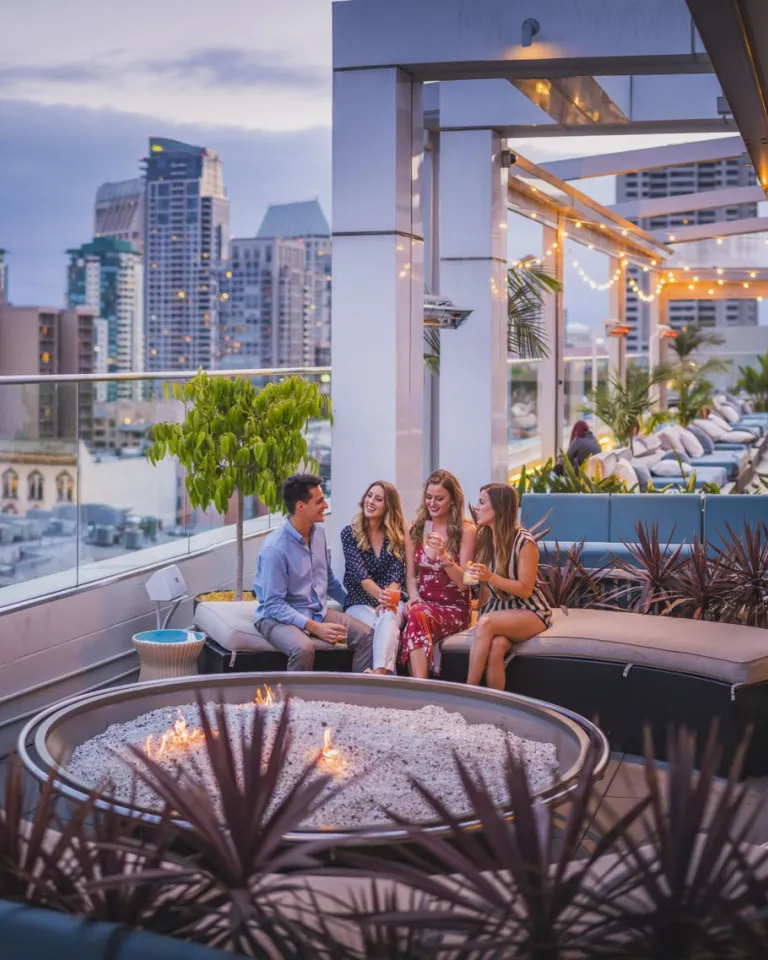 Four people sit around a fire pit on a rooftop lounge at sunset, surrounded by modern buildings and string lights, with city views in the background.