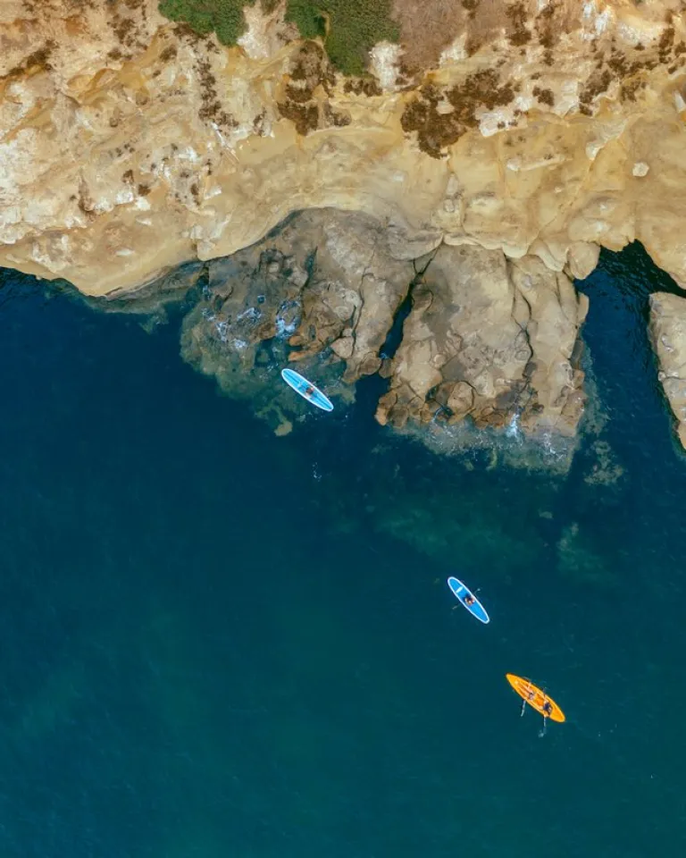 Aerial view of three paddleboards, one orange and two blue, floating near rocky cliffs in clear blue water.