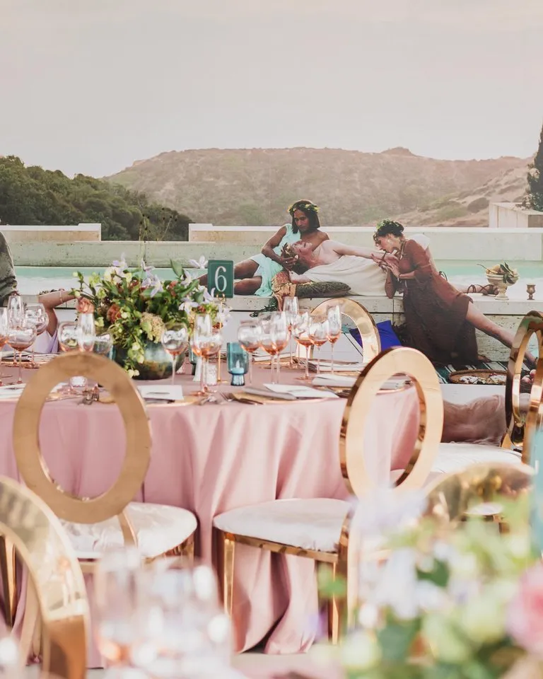 Outdoor event with a round table set for dining in the foreground; people in costumes recline on steps by a pool in the background with greenery and hills visible.
