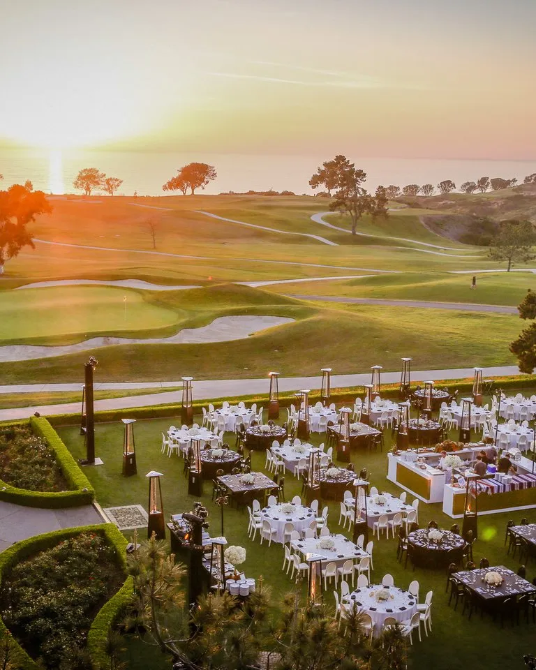 Outdoor event setup with round tables and chairs arranged on a lawn next to a golf course at sunset, with trees and a glowing sky in the background.