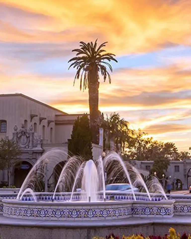 A fountain surrounded by flowers in front of historic Spanish-style buildings at sunset, with a tall tower and palm trees in the background.
