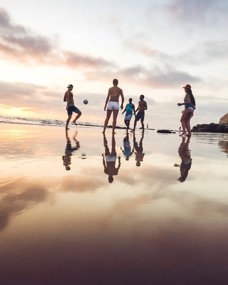 A group of people stand and play with a ball on a wet sandy beach at sunset, with cliffs and ocean visible in the background.