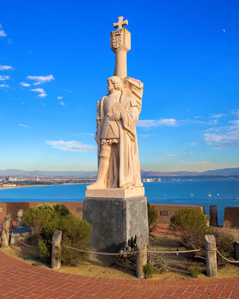 A stone statue of a historical figure stands on a raised platform overlooking the ocean, with people observing the coastal view and city skyline in the background.