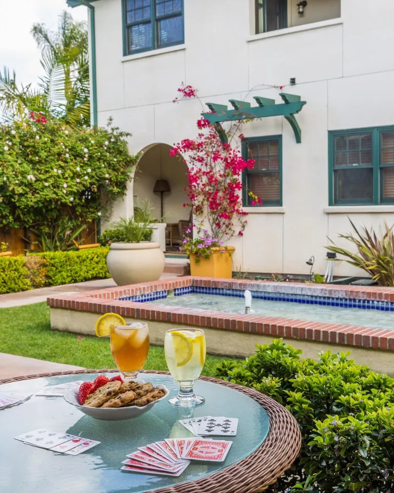 A round wicker table with playing cards, cookies, drinks, and fruit sits on a patio overlooking a small pool and garden next to a white building.