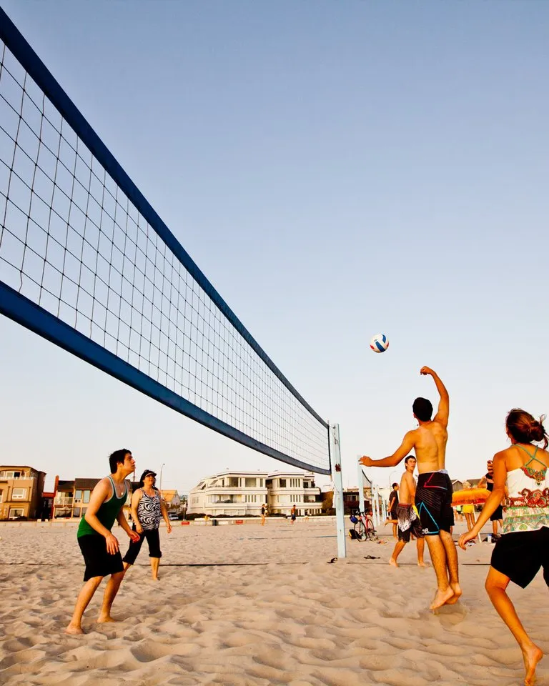 A group of people play beach volleyball on sand near the ocean, with houses and palm trees visible in the background under a clear sky.