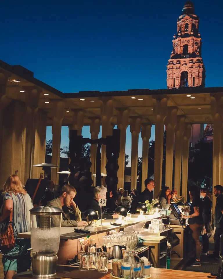 People gather at an outdoor bar and dining area in the evening; a tall illuminated tower is visible in the background against a dark blue sky.