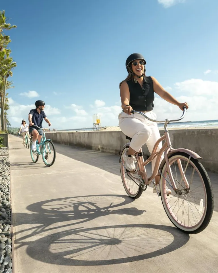 Two people ride bicycles along a beachside path on a sunny day, with palm trees, buildings, and the ocean visible in the background.
