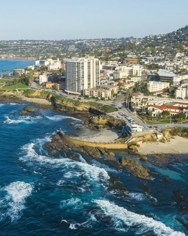 A coastal city with buildings along the shoreline, rocky cliffs, and waves crashing onto the beach under a clear blue sky.