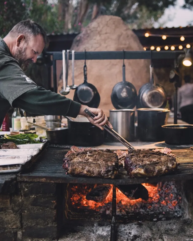 A man in a chef’s coat grills large pieces of meat over an open flame in an outdoor kitchen with hanging pots and string lights.