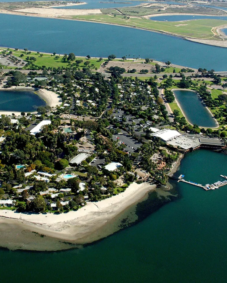 Aerial view of a small, lush island connected by a bridge, surrounded by water with sandy shores and greenery, featuring buildings and a dock.
