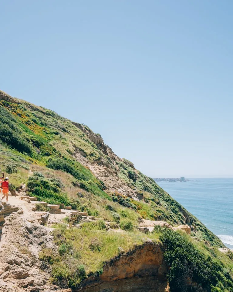 A group of people walk along a narrow cliffside path overlooking the ocean on a sunny day, with green hills and blue sky in the background.