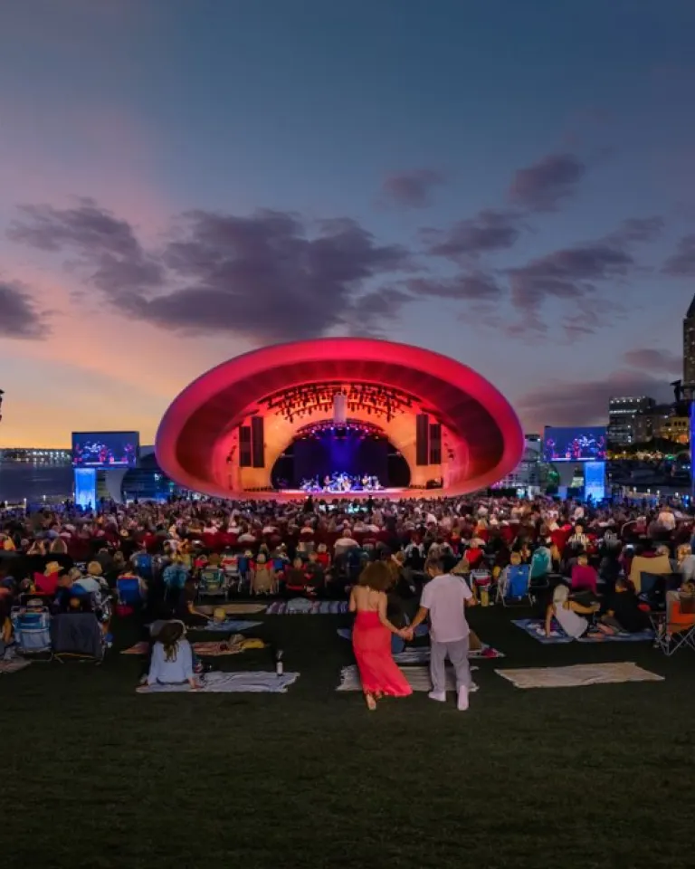 The Shell. A large crowd sits on a lawn facing an outdoor concert stage with a red-lit arch, city skyscrapers, and a sunset sky in the background.