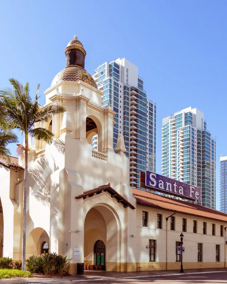 Historic Santa Fe Depot with Spanish-style architecture in front of modern high-rise buildings and palm trees under a clear blue sky.