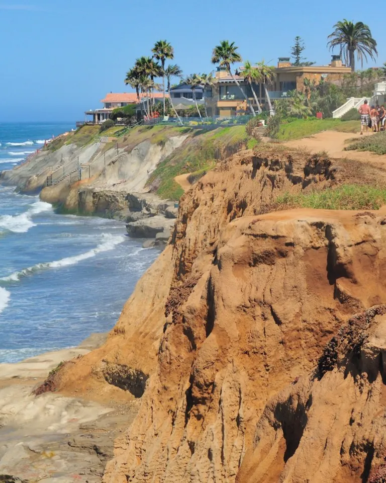 People walk along a sandy cliffside trail overlooking the ocean, with houses and palm trees in the background on a clear day.