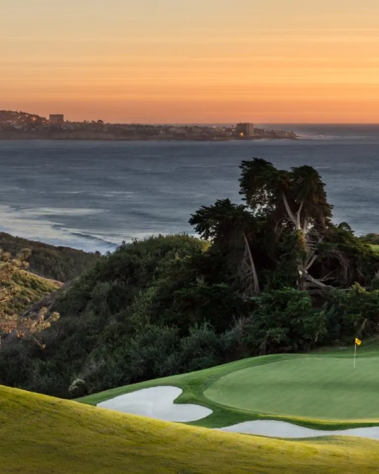 Torrey Pines North Golf Course 15 green with sand bunkers overlooks the ocean at sunset, with distant hills and a coastal town visible in the background.