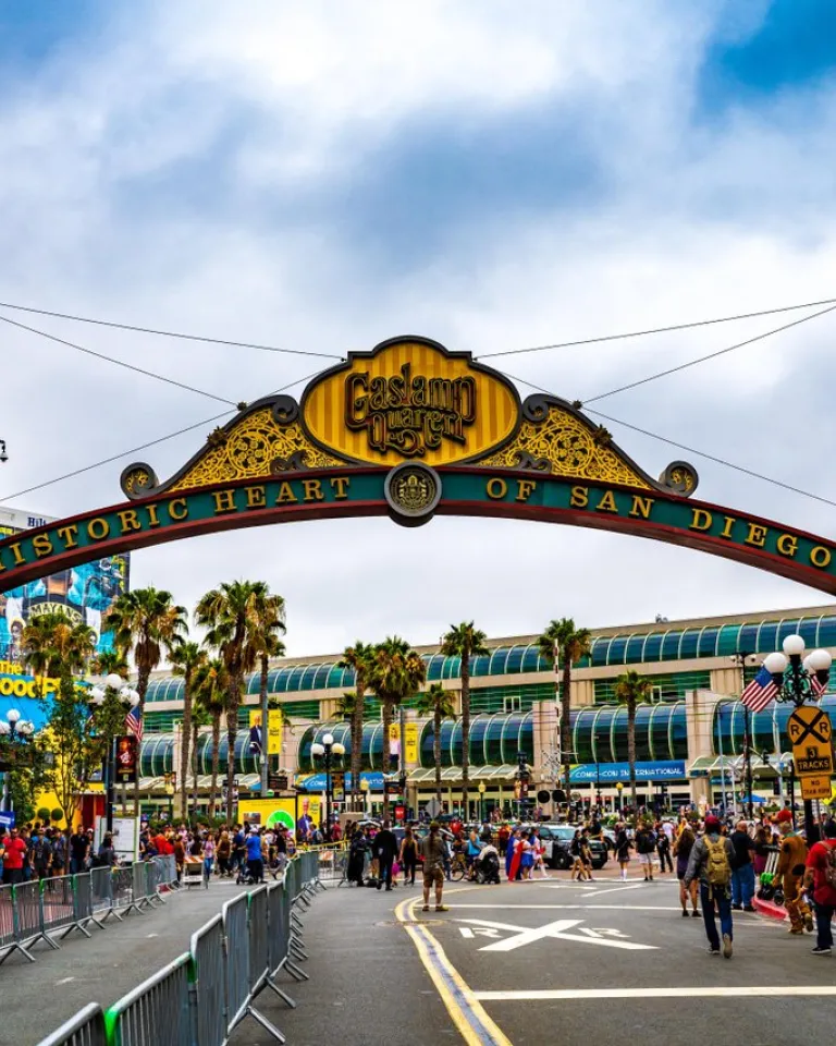 A large archway reading "Historic Heart of San Diego" stands over a busy street filled with people, with palm trees and buildings visible in the background, in San Diego's historic Gaslamp Quarter.
