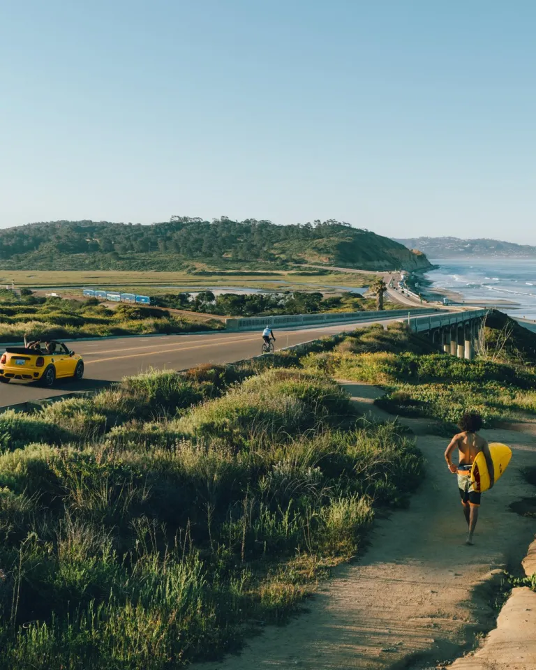 A person carries a surfboard along a path toward the beach as a yellow convertible drives on a coastal road under a clear sky.