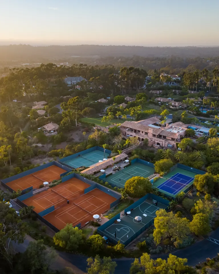 Aerial view of a sports complex with multiple tennis courts, a basketball court, nearby buildings, and lush greenery at sunset over Rancho Santa Fe in San Diego County.