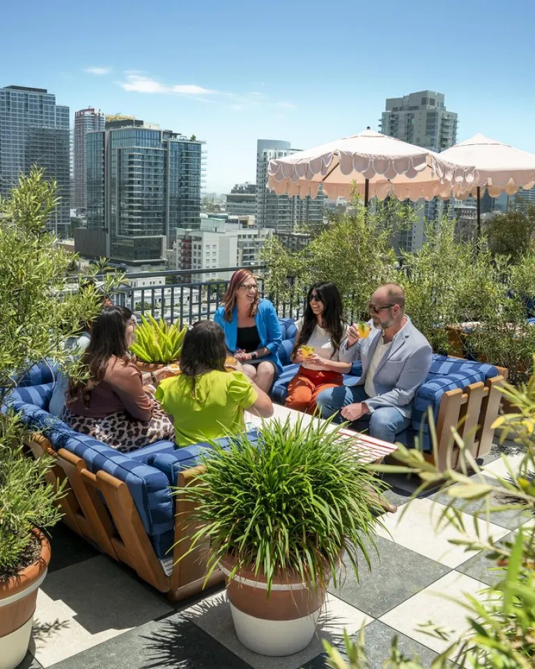 Five people sit and talk on a rooftop patio surrounded by plants, with San Diego city buildings in the background and a pink umbrella providing shade.