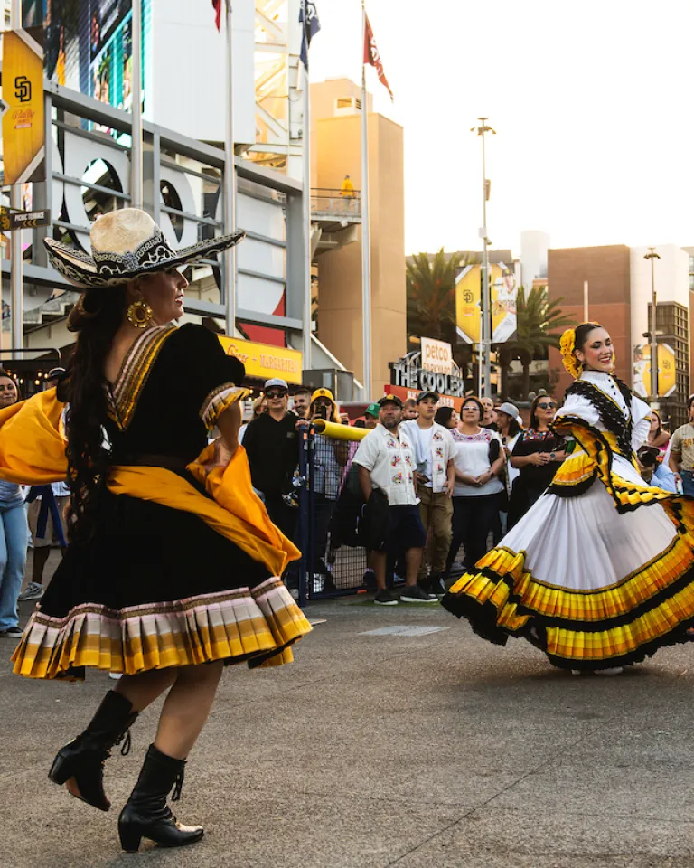 Two folklórico dancers perform in front of a crowd outside Petco Park in San Diego. The dancer in the foreground wears a black dress with yellow accents and a wide-brimmed sombrero, twirling a yellow shawl, while the dancer behind her wears a flowing white, yellow, and black ruffled dress, holding out the skirt as she spins. Baseball fans in jerseys and caps watch from behind a barrier, with stadium signage, flags, and downtown buildings visible in the background at sunset.