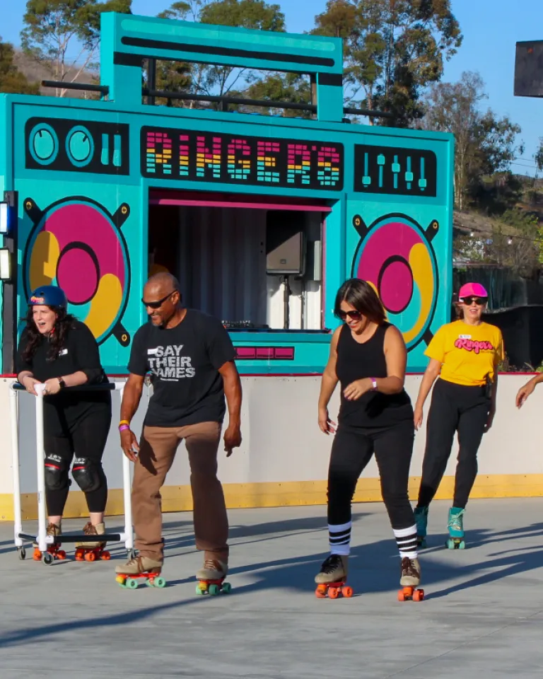 Five adults roller skating side by side at an outdoor rink at Ringers Roller Rink in San Diego’s Encanto neighborhood, all smiling and mid-glide in the sun. Behind them is a brightly colored backdrop designed like a giant boombox with the word “Ringers” on it, framed by trees and blue sky.