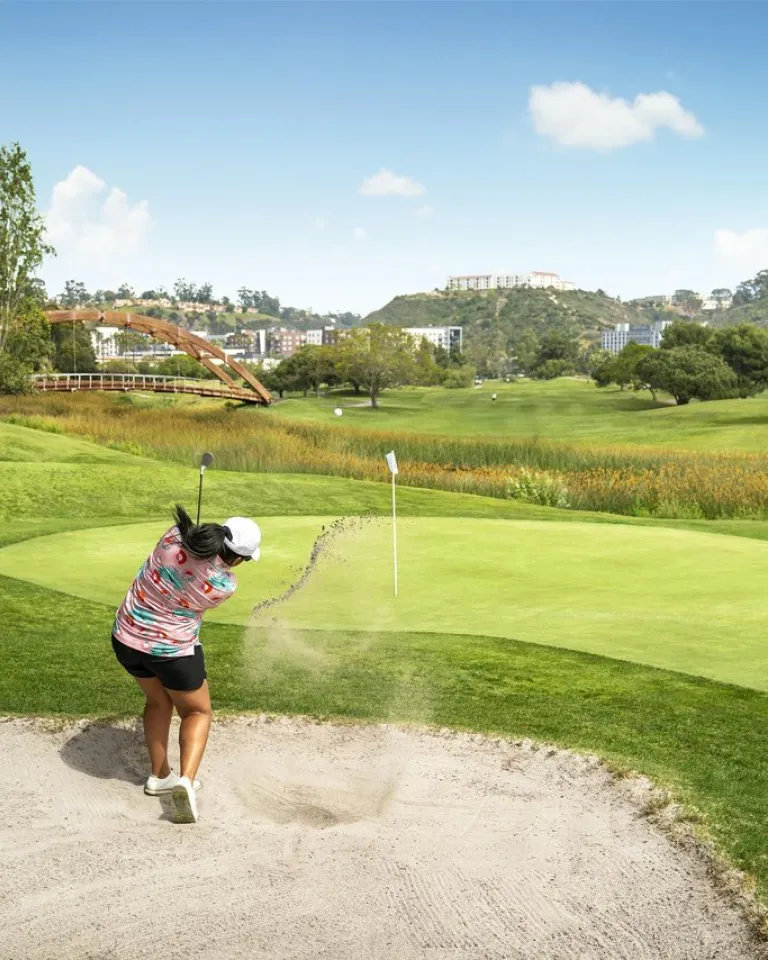 A golfer in shorts and a pink shirt drives a golf ball out of a sand trap toward the green on a sunny day in San Diego's beautiful Mission Valley.