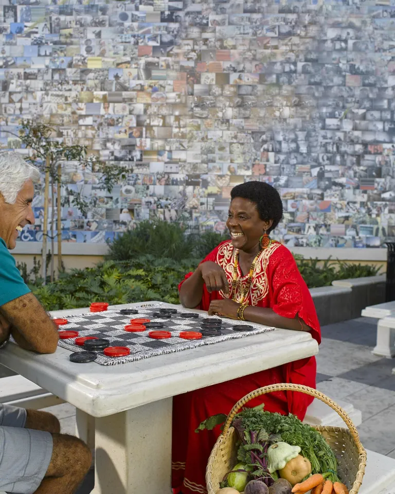 Two people sit across from each other playing a game with large red and black checkers on a stone table in an outdoor plaza in City Heights, San Diego, California. The person on the left wears a teal shirt and khaki shorts; the person on the right wears a bright red dress with gold embroidery and is smiling. A large mosaic-style mural made of many small photos fills the wall behind them, and a basket of fresh vegetables and herbs rests on the ground beside the table.